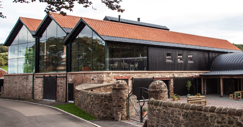 Modern glass building with a slate roof, blending contemporary design with natural surroundings, nestled in green landscaping.
