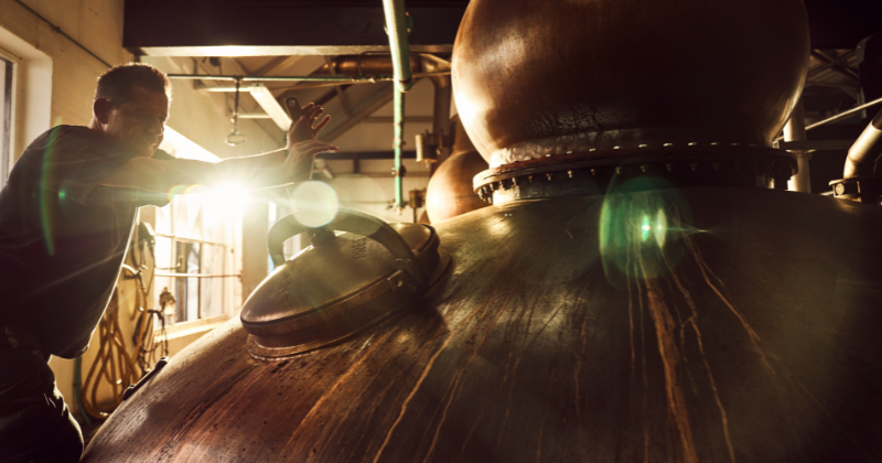 A distiller adjusts equipment in a warm, industrial setting, with sunlight streaming in, highlighting the brewing process.