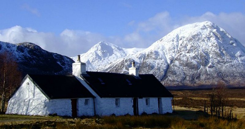 A picturesque white cottage in a serene landscape, surrounded by snow-capped mountains under a bright blue sky.