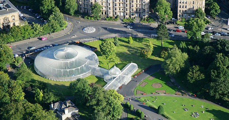 Aerial view of a botanical garden with glass structures surrounded by lush greenery and a road, showcasing a scenic urban setting.
