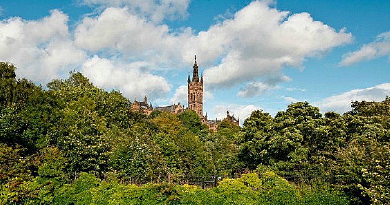 A historic tower rises above lush greenery and trees under a bright blue sky with fluffy clouds.