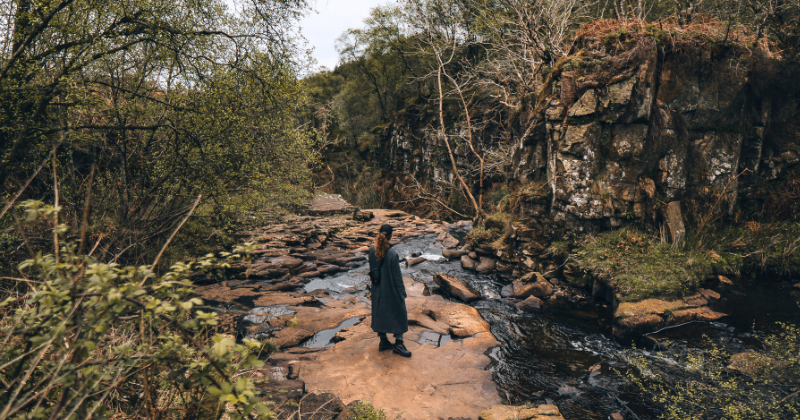 A person stands by a serene river surrounded by lush greenery and rocky cliffs, enjoying the natural beauty of the UK.