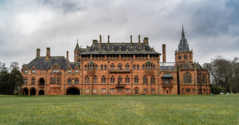 Historic red-brick mansion with ornate architecture, surrounded by a grassy landscape under a cloudy sky.