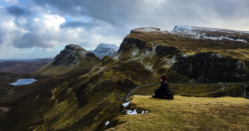 A person sits peacefully on a grassy hilltop, overlooking dramatic mountains and a serene lake under a cloudy sky.