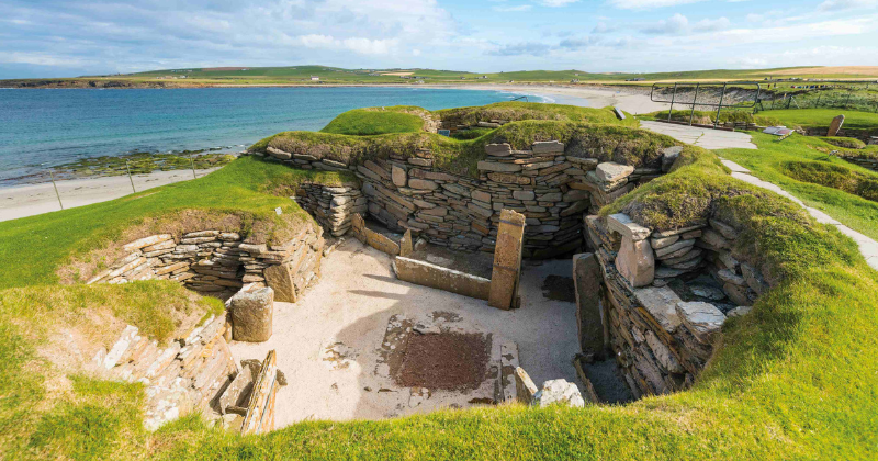 Ancient stone ruins with grassy mounds overlook a serene beach and coastline, showcasing historic architecture by the water.