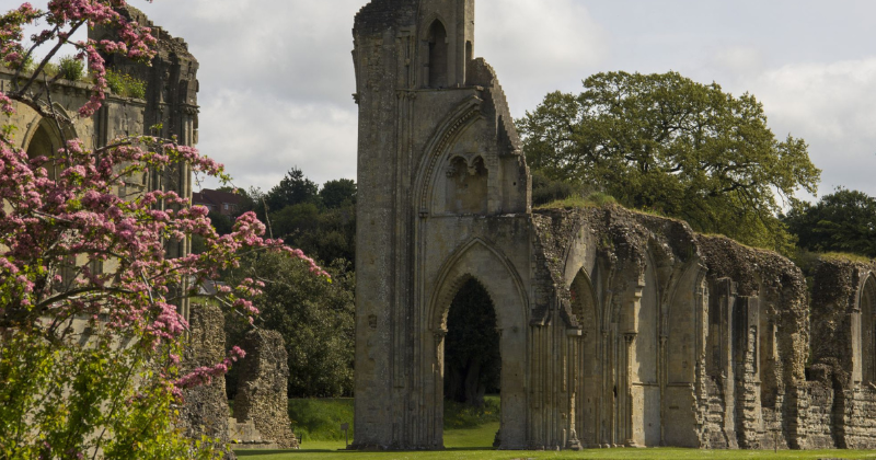Ruins of an ancient stone structure surrounded by lush greenery and flowering plants under a cloudy sky.
