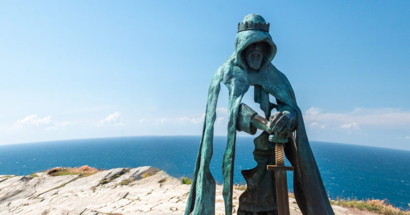 A bronze statue of a robed figure holding a sword, set against a blue ocean and clear sky backdrop.