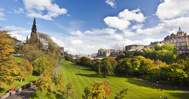 A scenic view of a park in Edinburgh, featuring lush greenery, people enjoying the space, and historic buildings in the background.
