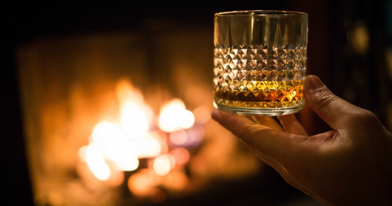 a hand holding a glass of whisky before a fireplace