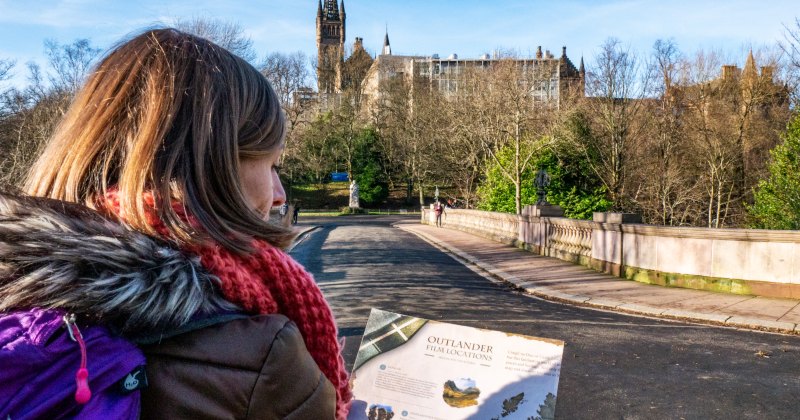A woman with a map stands on a bridge, overlooking a park with trees and a historic building in the background.