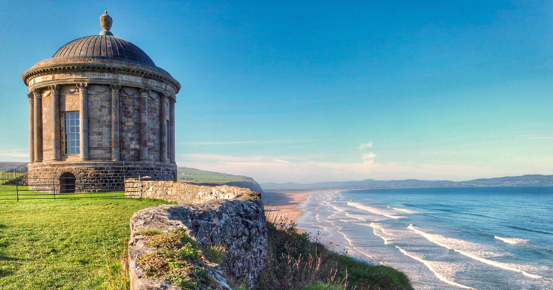 A scenic view of a historic round building overlooking a sandy beach and the ocean under a clear blue sky.