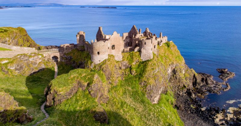 Ruins of a historic castle perched on a cliff, overlooking the ocean, surrounded by lush green landscapes.
