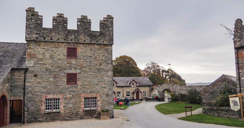 Historic stone buildings with a castle-like structure, set along a winding road, with people and greenery in the surroundings.