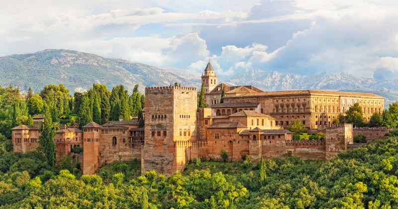 Scenic view of the Alhambra palace surrounded by lush greenery and mountains under a cloudy sky.