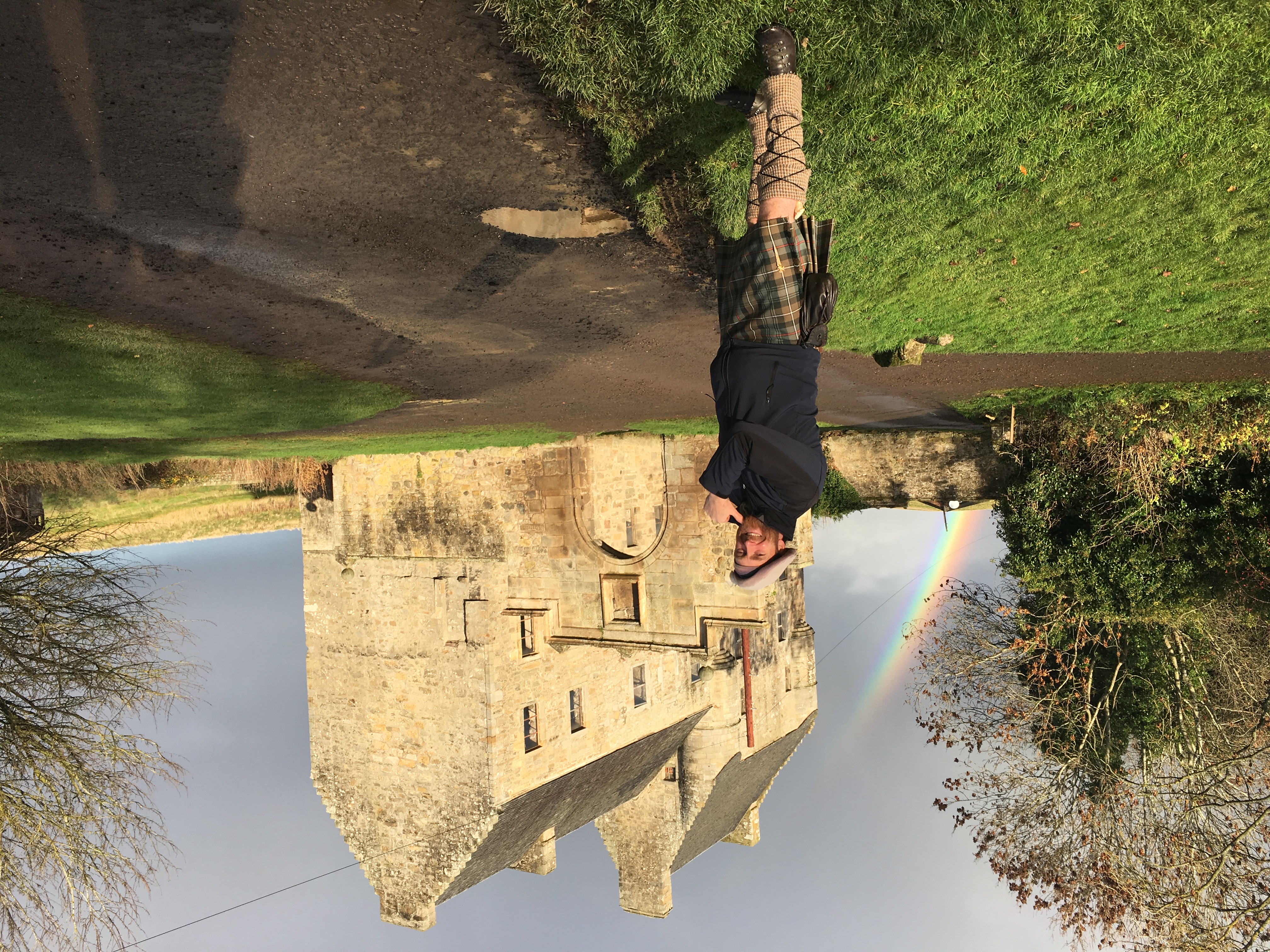 A man in a kilt poses beside a historic stone building, with a rainbow appearing in the background against a cloudy sky.