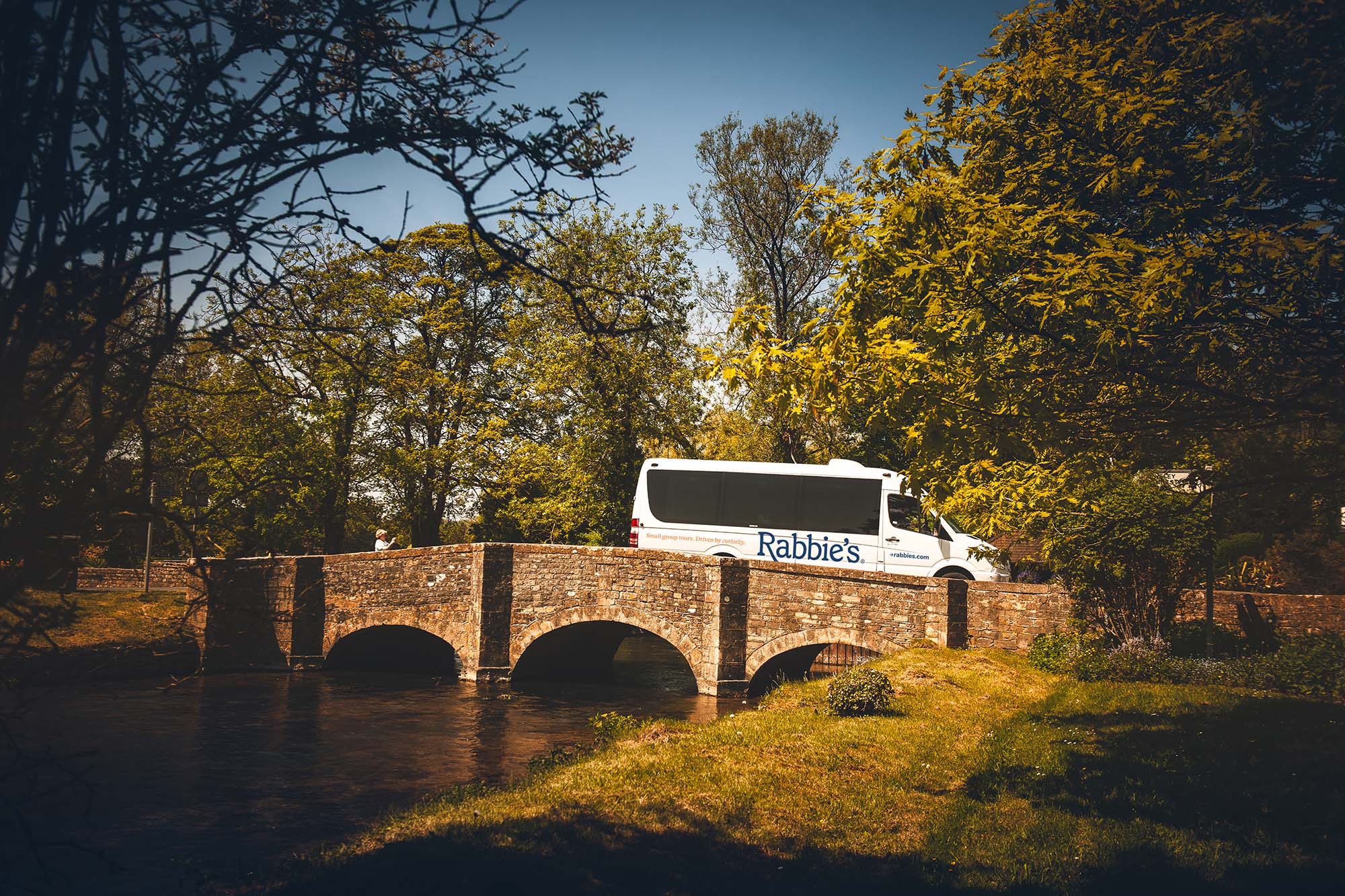 A Rabbie's Tours van crosses a picturesque stone bridge surrounded by lush greenery and a serene waterway.