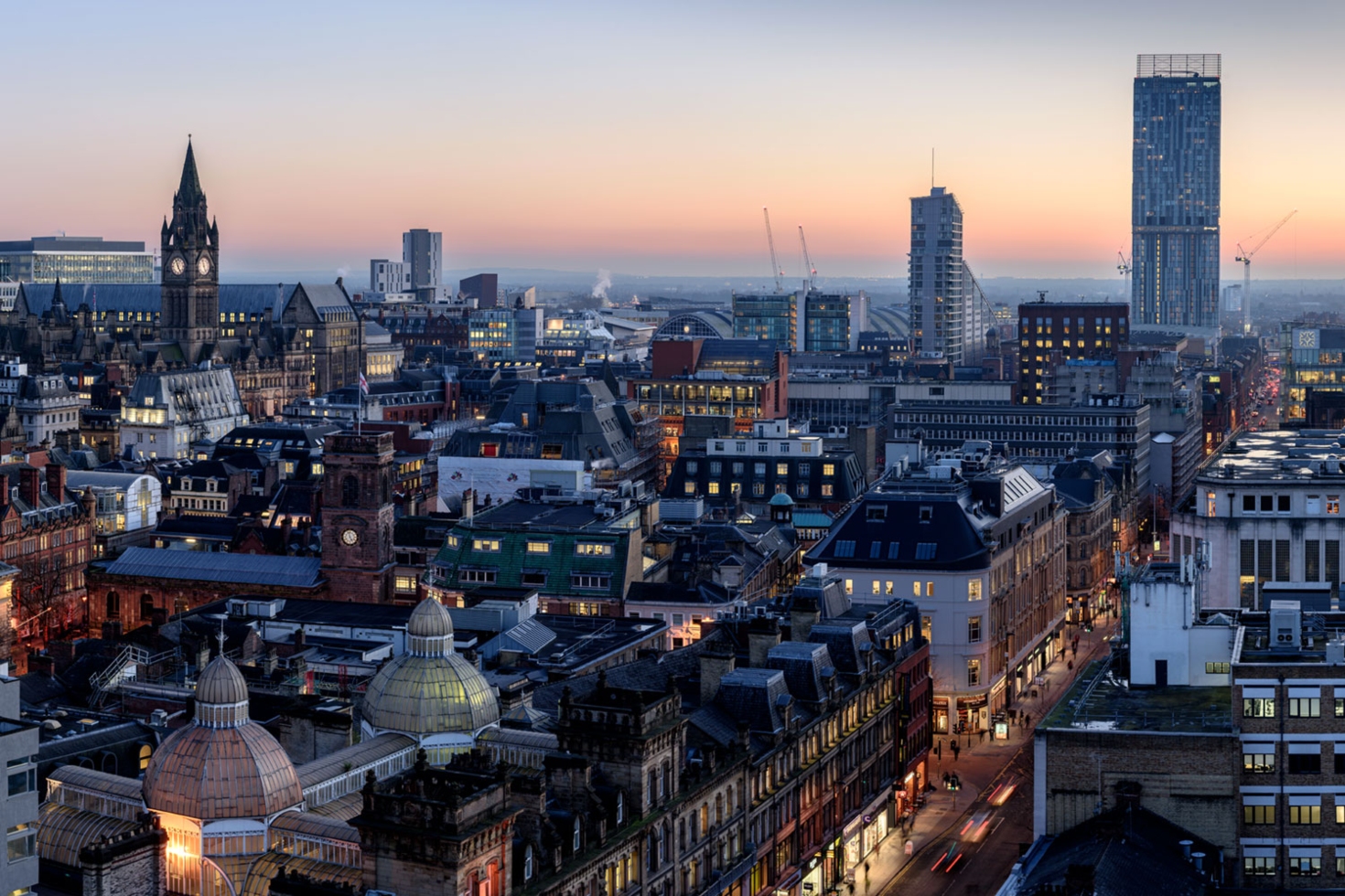 Cityscape of Manchester at dusk, showcasing historic architecture and modern buildings against a colorful sky.