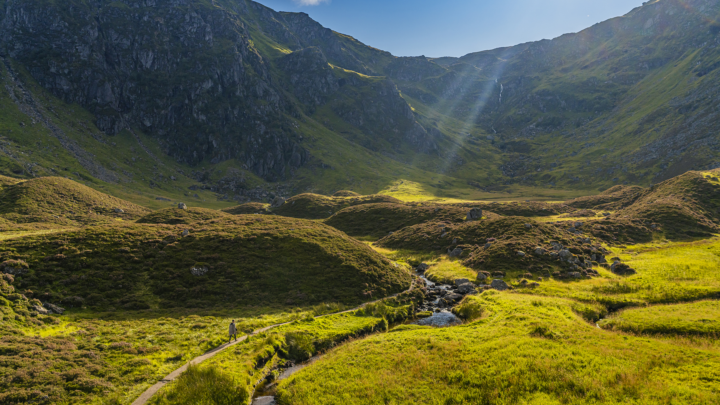 A serene landscape of rolling green hills and a winding path, illuminated by sunlight streaming through the mountains.
