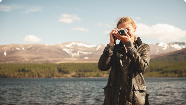 A person with a camera captures the scenic beauty of a lake surrounded by mountains under a clear blue sky.