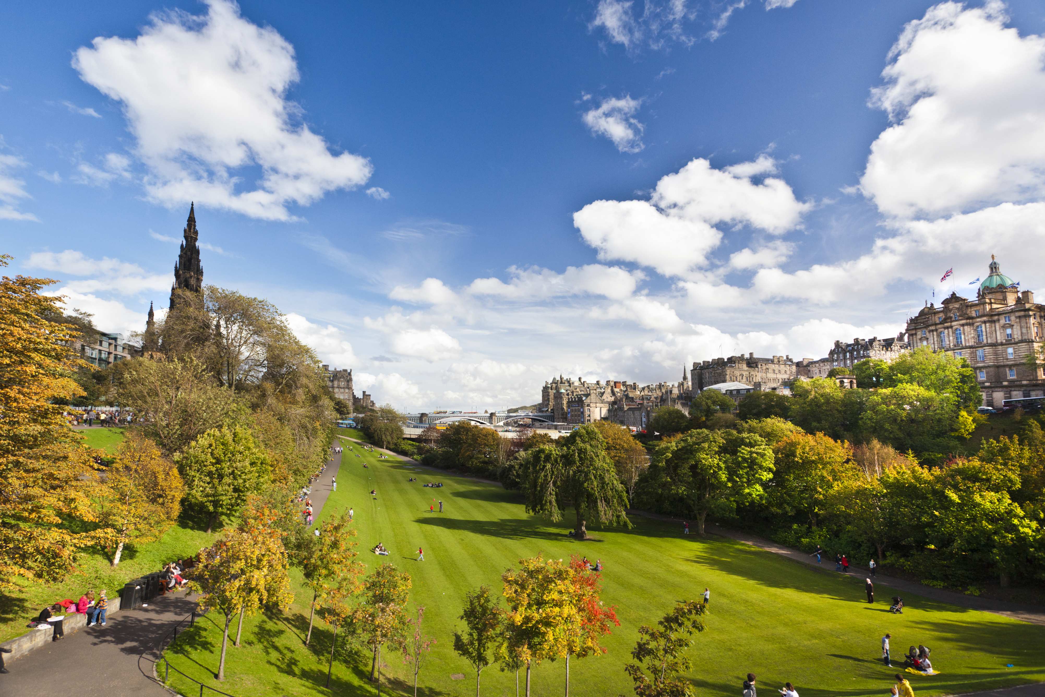 A vibrant park in Edinburgh with green lawns, colorful trees, and a backdrop of historical buildings under a bright blue sky.