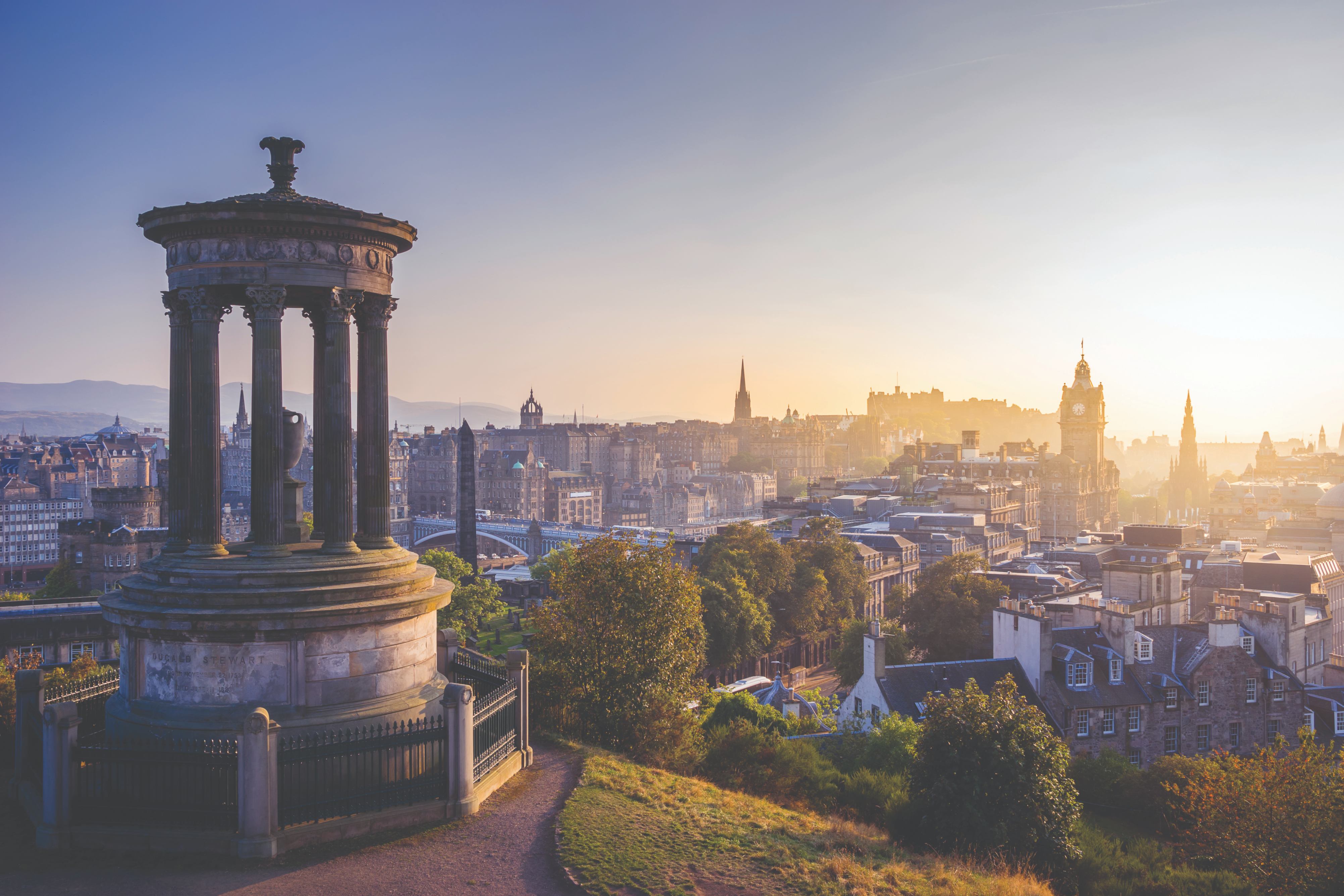 Sunrise over Edinburgh, featuring a historical monument with a panoramic view of the city skyline and soft morning light.