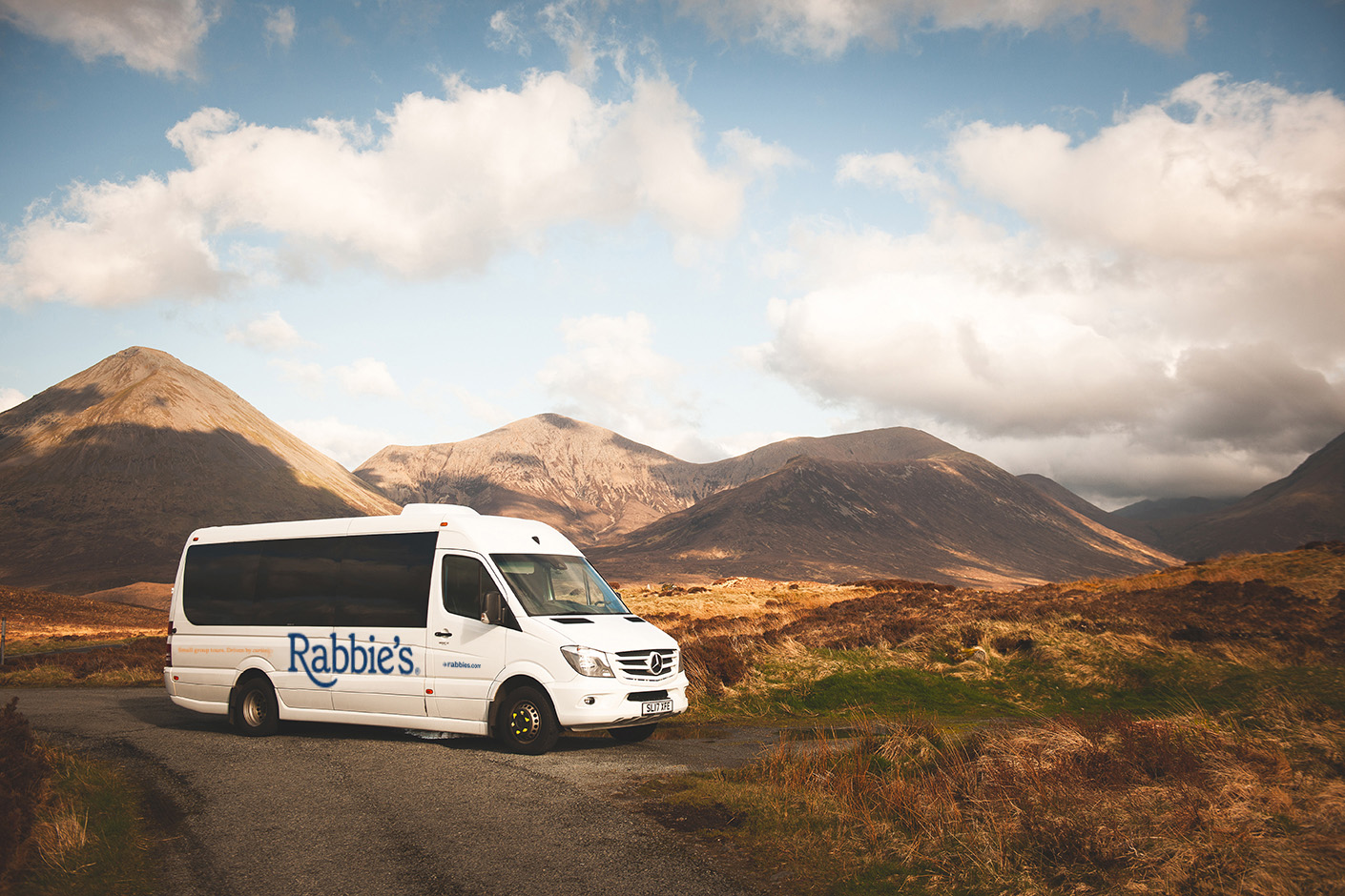 A Rabbie's Tours minibus parked on a scenic road, surrounded by mountains and open landscape under a cloudy sky.