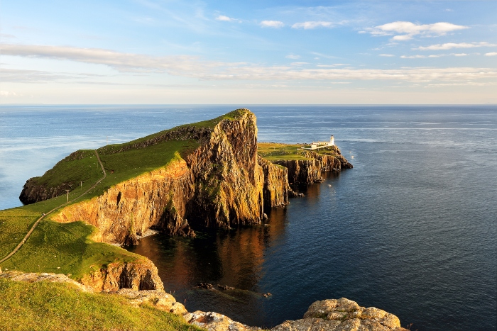 Stunning coastal view of a rocky cliff by the sea, featuring lush greenery and a distant lighthouse under a clear blue sky.