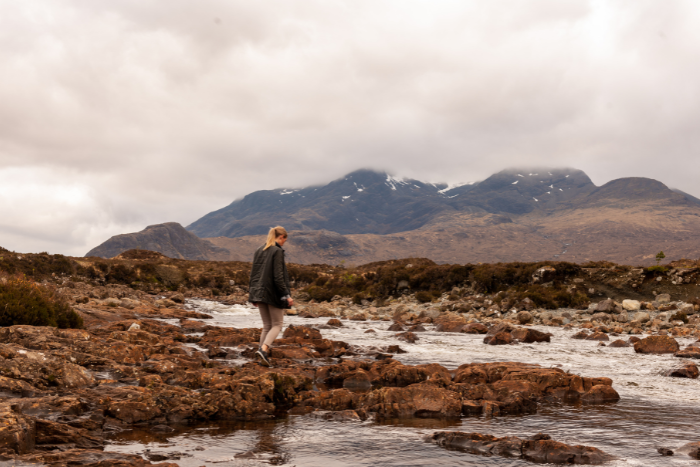 A woman walking along rocky terrain by a stream, with mountains and cloudy skies in the background, showcasing the UK's natural beauty.