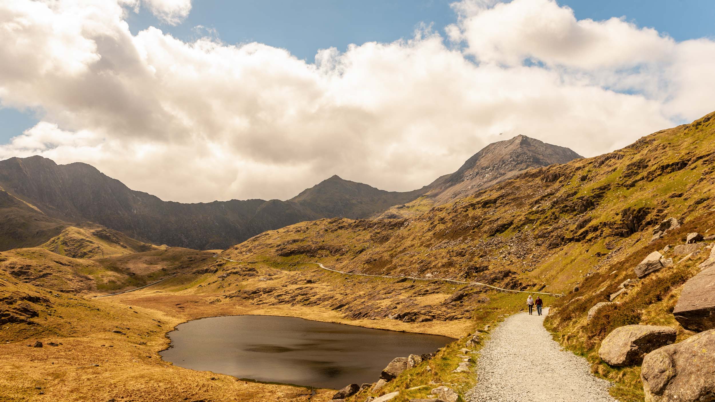 Scenic landscape featuring a winding path, mountains, and a serene lake under a partly cloudy sky in the UK.